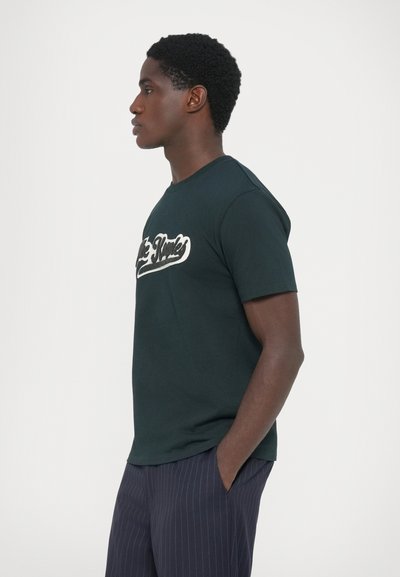 Tumši zaļš īsām piedurknēm T-krekliņš ar baltu druk�ātu logo uz priekšpuses, apaļā kakla izgriezumā, izgatavots no mīksta auduma, kombinēts ar svītrainām biksēm.