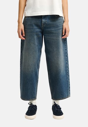 Personne portant un jean en denim bleu à jambes larges, des chaussettes blanches, des baskets noires et une chemise blanche, debout devant un fond clair.