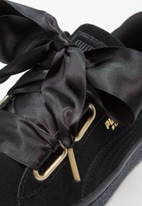 Gros plan sur une chaussure Puma noire avec des lacets larges en ruban de satin noués en boucle, texture en daim et œillets dorés sur fond blanc.