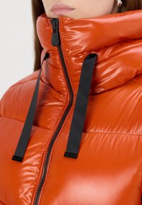 Orange puffer jacket with a high collar, glossy finish, black zipper, and adjustable black drawstrings. Features quilted texture.