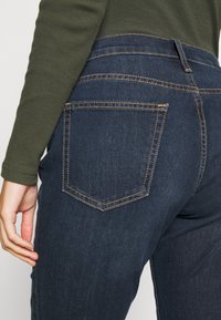Gros plan d'une personne portant un jean en denim bleu foncé et un haut vert à manches longues, montrant la poche arrière et une partie de sa main.