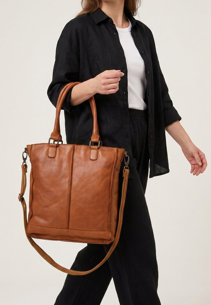 Femme vêtue d'une chemise noire et d'un pantalon avec un haut blanc, portant un grand sac cabas en cuir marron par les poignées.