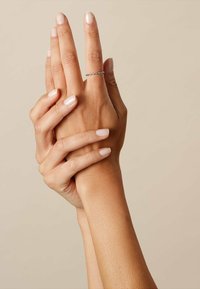 Silver ring med en vriden design på en hand, med släta, välvårdade fingrar och ljusbeige nagellack, händer sammanflätade över varandra.