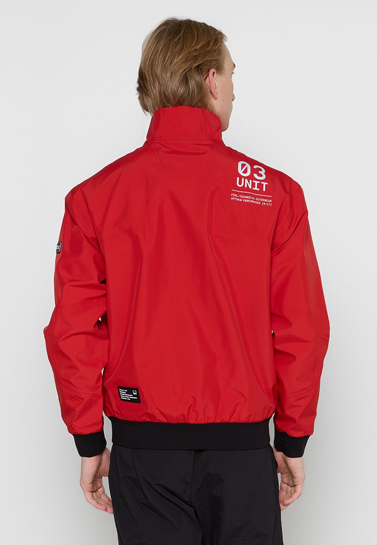 Chaqueta de nailon roja con un alto cuello, puños elásticos y cinturilla. Presenta texto impreso en blanco y un bolsillo en el pecho izquierdo. Textura suave.