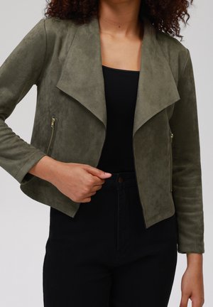 Femme portant une veste courte en daim vert olive avec de larges revers et des poches zippées, par-dessus un haut noir et un pantalon.