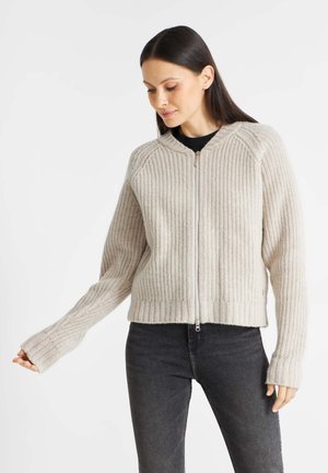 Beige knitted sweater with a zip front, ribbed texture, and contrasting fuller sleeves. Worn over a black top and paired with dark jeans.