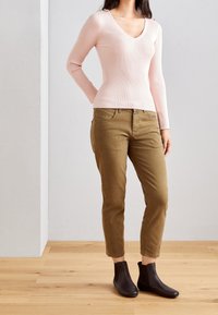 Donna che indossa un maglione a collo a V ribbed rosa chiaro, pantaloni marroni a sigaretta e stivaletti neri, in piedi su un pavimento di legno contro un muro bianco.