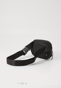 Svart stoffbelteveske med glidelås på toppen, justerbar rem og plastspenne. Har en glatt tekstur og minimalistisk design.