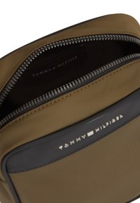 Καφέ νάιλον τσάντα με μαύρο περίγραμμα, ασημένιο φερμουάρ μερικώς ανοιχτό και το λογότυπο "Tommy Hilfiger" σε ασημί στην μπροστινή πλευρά.