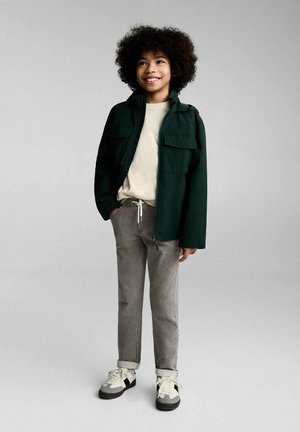 Giacca a zip verde scuro con due tasche sul petto, felpa beige chiaro, pantaloni grigi e sneakers nere con accenti bianchi.