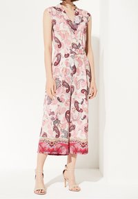 Mono sin mangas de color rosa con patrón de paisley, cintura ceñida y piernas amplias. Presenta un cuello y un dobladillo texturizado en rosa oscuro. Las sandalias son en tono nude.