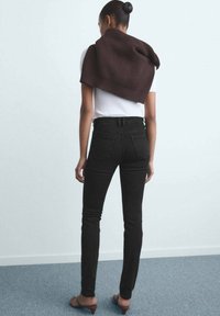Vaqueros negros de cintura alta con un diseño ajustado, combinados con una camiseta blanca de canalé y un suéter de punto marrón colocado sobre los hombros.