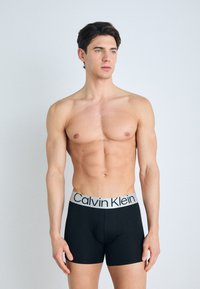 Fekete boxer alsónadrág elasztikus anyagból, szürke derékrésszel, amelyen a "Calvin Klein" logó szerepel. A modellt a testhez simuló dizájn jellemzi.