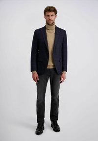 Mann in dunkel kariertem Blazer, beigem Rollkragenpullover, schwarzen Jeans und schwarzen Lederschuhen, steht vor einem einfarbigen Hintergrund.