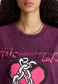 Camiseta de color púrpura oscuro con un diseño gráfico que presenta un corazón rosa y un detalle de cadena blanca. El texto "THIS LOVE" está impreso en letras negras en negrita.