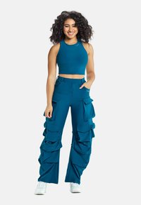 Top corto teal senza maniche abbinato a pantaloni cargo teal con tasche a strati e volant. Sneakers bianche completano l'outfit.