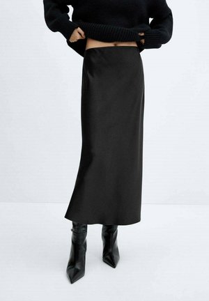 Jupe midi en satin noir avec une texture lisse et une silhouette droite, portée avec un pull noir en maille ample et des bottes noires pointues.