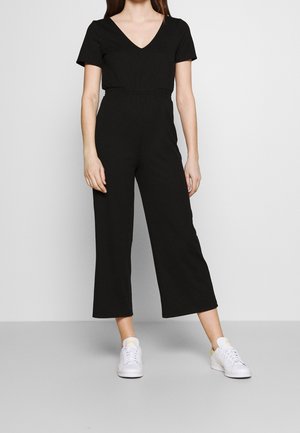 Czarny prążkowany kombinezon z krótkimi rękawami, dekoltem w serek, zaznaczoną talią i szerokimi nogawkami, w połączeniu z białymi trampkami.