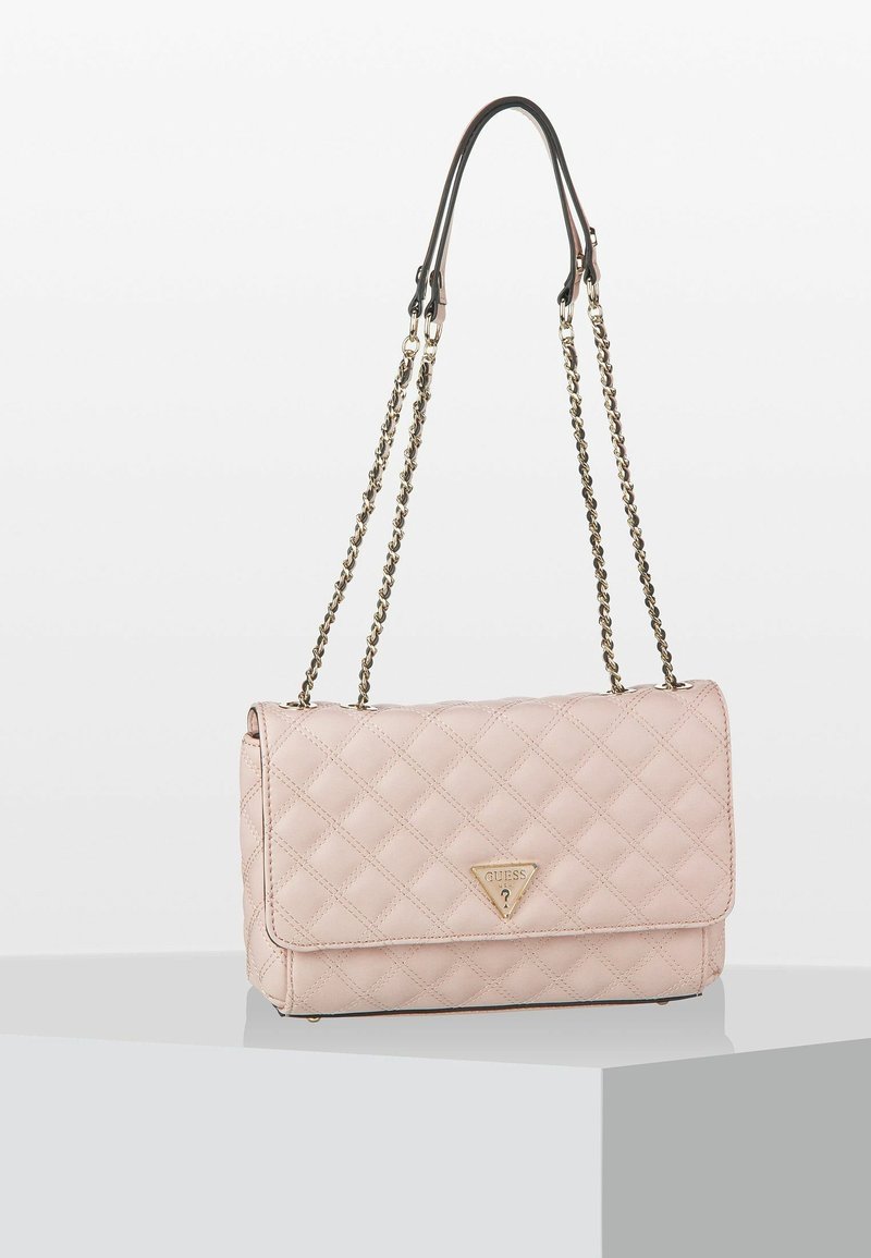 Sac à bandoulière matelassé rose clair avec des sangles en chaîne dorée et un logo triangulaire Guess sur le rabat avant, posé sur une surface blanche.