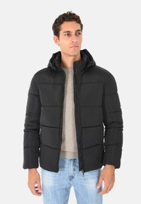 Giubbotto imbottito nero con cappuccio, design trapuntato, zip frontale, tasche laterali e polsini elasticizzati, indossato sopra un maglione grigio chiaro.