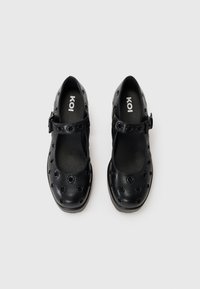 Black Mary Jane shoes with large circular cutouts and a secure buckle strap. Smooth texture, rounded toe, subtle cushioned insole.