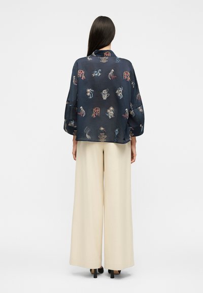 Femme aux longs cheveux foncés portant un chemisier bleu marine à imprimés abstraits et un pantalon large crème, debout de dos sur un fond blanc.
