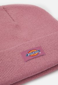 Ružový pletený čiapka s prehnutým okrajom, ktorá obsahuje obdlžníkovú nášivku s logom "Dickies" v modrých, červených a žltých farbách.