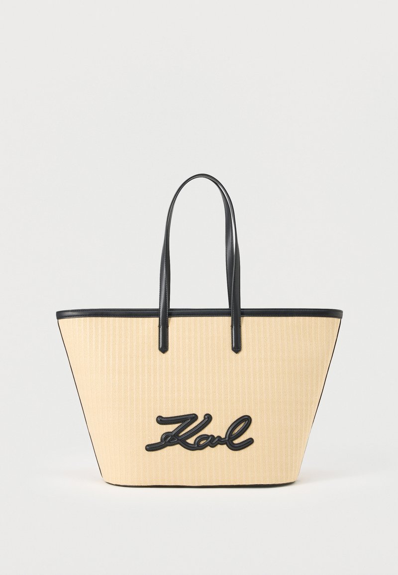 Μπεζ πλεκτή τσάντα ώμου με μαύρες δερμάτινες λαβές και περιθώριο, με μαύρο καλλιγραφικό λογότυπο «Karl» στο μπροστινό μέρος.