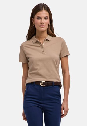 Vrouw draagt een beige polo met korte mouwen, ingestopt in marineblauwe broek met een bruine riem, staand tegen een effen achtergrond.