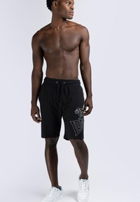 Shorts en coton noirs avec impression de logo, taille avec cordon de serrage, design longueur genou, assortis à des baskets blanches. Le modèle se tient de profil.