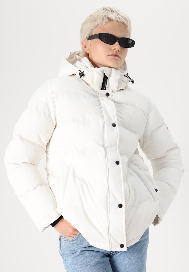 Weißer Pufferjacke mit gestepptem Design, versehen mit schwarzen Druckknöpfen und einer Kapuze mit Kordelzug, getragen über Denim. Glatte Textur.