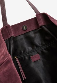 Bordeaux ruskinds tote bag med sort stofforing, der har en tryklås og en indvendig lynlåslomme med et lædermærke.
