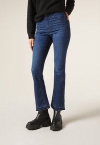 Jeans évasés bleu nuit en denim, avec une taille haute, un ourlet brut et une coupe près des hanches, associés à des bottines noires à semelle épaisse.