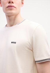 T-shirt beige à manches courtes en tissu doux. Présente un col rond avec des rayures noires et blanches, et un petit logo noir "BOSS" sur la poitrine.