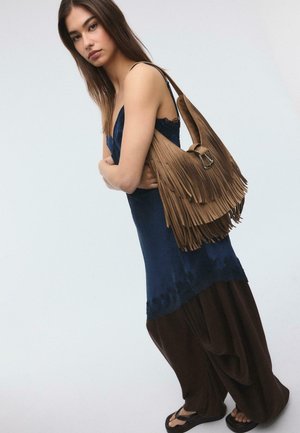 Junge Frau in einem dunkelblauen ärmellosen Kleid und lockeren braunen Hosen, die eine große braune Schultertasche mit Fransen trägt, steht vor einem einfarbigen Hintergrund.