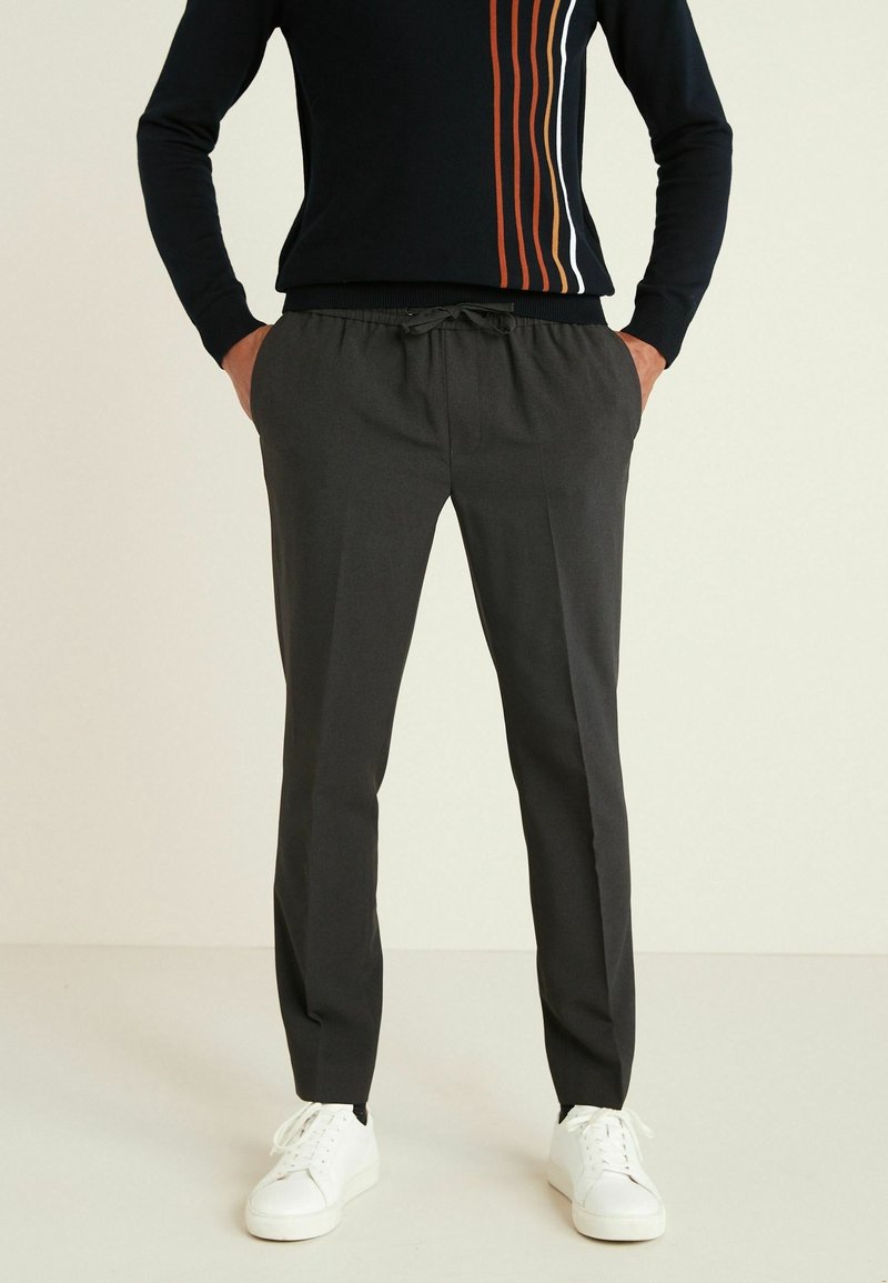 Personne portant un pantalon gris foncé à cordon, un pull noir avec des rayures verticales orange et blanches, et des baskets blanches, debout les mains dans les poches.