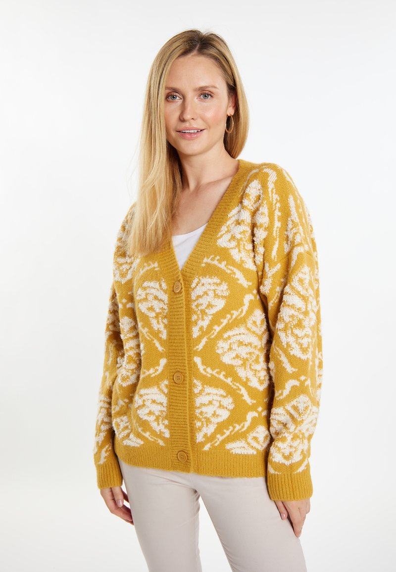 Cardigan giallo senape con morbidi motivi floreali bianchi, polsini e orlo a coste, e scollatura a V con chiusure a bottone sul davanti.