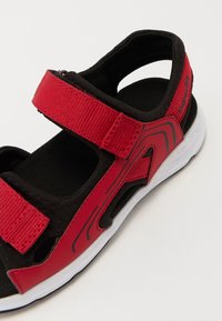 Røde og sorte sandaler med justerbare stropper, en teksturert fotseng og en hvit såle med en sort aksent på bunnen.