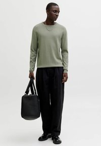 Pull vert clair à manches longues, coupe décontractée, associé à un pantalon noir à jambes larges et des chaussures en cuir noir, tenant un sac noir.
