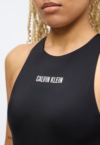 Κοντινό πλάνο ενός ατόμου που φοράει μαύρο αμάνικο μπλουζάκι με την επιγραφή "CALVIN KLEIN" σε λευκό στο στήθος.
