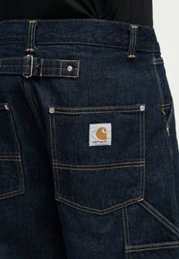 ciemnoniebieskie spodnie jeansowe z widocznym żółtym przeszyciem, tylną kieszenią z prostokątną naszywką z logo Carhartt oraz regulowanym bocznym klamrą.