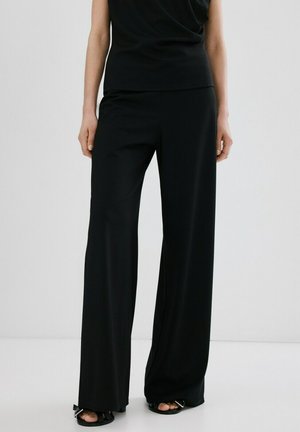 Kvinde iført sorte bukser med brede ben og sorte sandaler med åben tå, stående mod en ensfarvet lys baggrund.