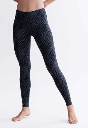 Person trägt schwarze Leggings mit hohem Bund und dezenten dunklen Zebramustern, steht barfuß mit leicht gespreizten Beinen vor einem einfarbigen Hintergrund.