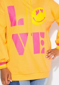 Felpa gialla brillante con il testo "LOVE" in bold rosa e una grafica di faccina sorridente. Presenta un cappuccio con coulisse e polsini a coste con strisce rosa e gialle.