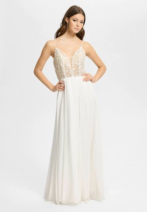 Junge Frau mit langem, dunklem Haar trägt ein weißes, ärmelloses Kleid mit besticktem Blumenmuster am Oberteil und einem fließenden Rock, posiert mit Händen in der Hüfte.