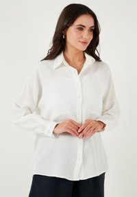 White button-up shirt featuring a textured fabric, pointed collar, and long sleeves, paired with navy pants and layered gold necklaces.