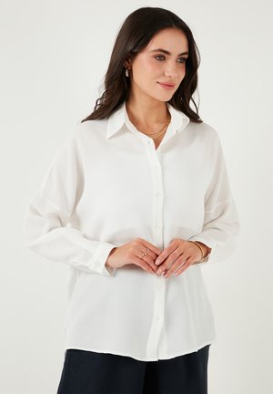 White button-up shirt featuring a textured fabric, pointed collar, and long sleeves, paired with navy pants and layered gold necklaces.