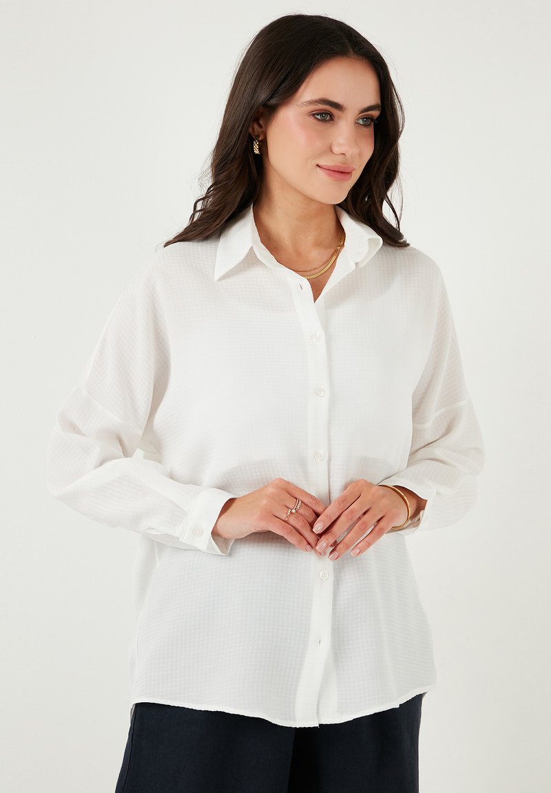 White button-up shirt featuring a textured fabric, pointed collar, and long sleeves, paired with navy pants and layered gold necklaces.