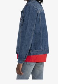 Chaqueta de mezclilla en azul medio con un corte clásico, cierre de botones y bolsillos laterales. Llevada sobre una camisa roja con dobladillo recto y jeans azules.