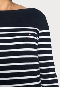 Pull bleu marine avec des rayures horizontales blanches et un petit logo rouge, blanc et bleu sur la poitrine, porté par une personne.